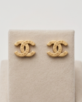 A pair of gold-tone Chanel logo stud earrings displayed on a beige textured jewelry stand against a light neutral background.